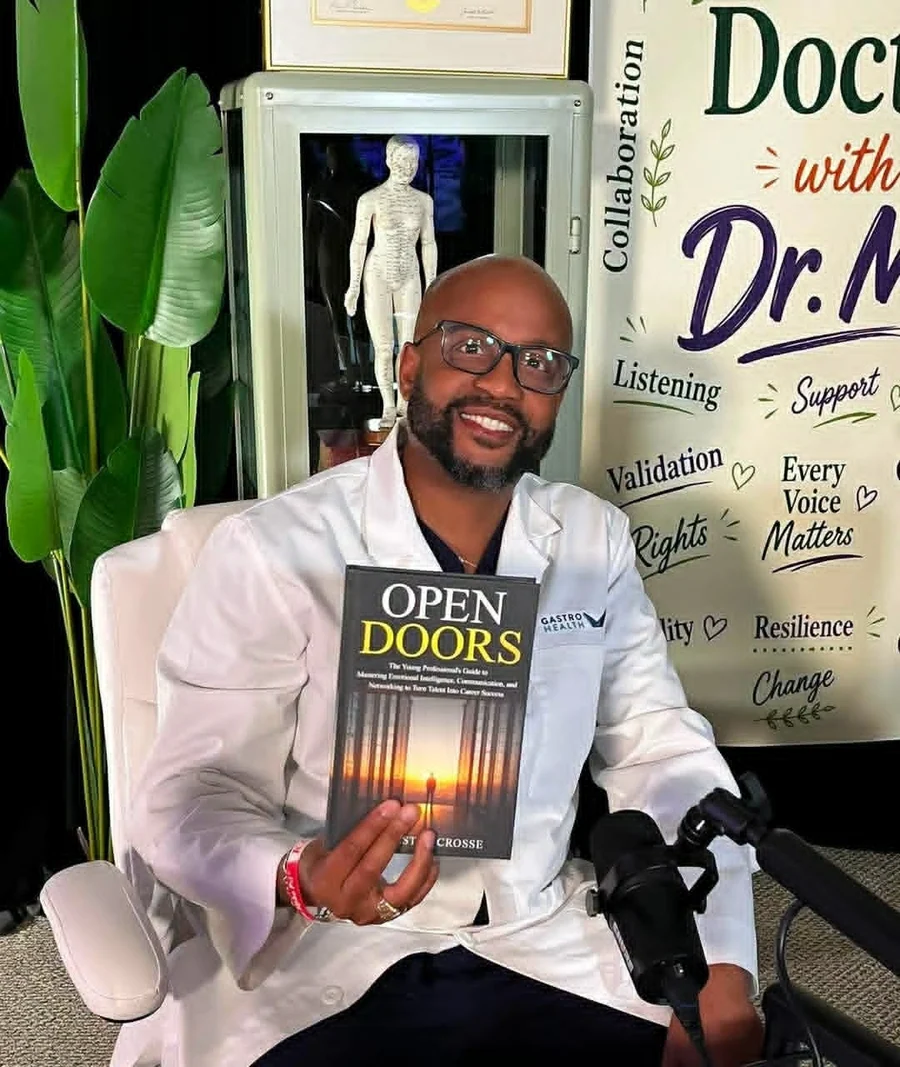 Dr. Kester Crosse in a white Gastro Health coat, seated on the set of Doctors with Dr. M, holding a copy of Open Doors. The show's values are painted on the backdrop behind him: collaboration, listening, support, validation, every voice matters, resilience, change.